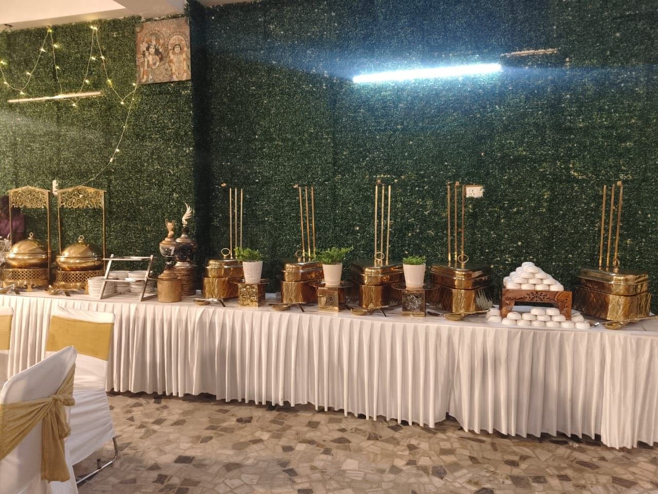 Full buffet counter of gold chafing dishes set against a green wall at a catered function