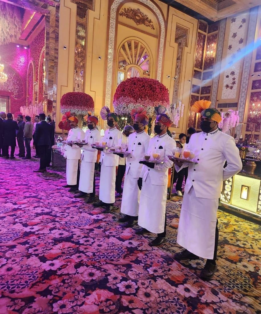 Service staff in white uniforms and coloured safas carrying welcome-drink trays at a wedding